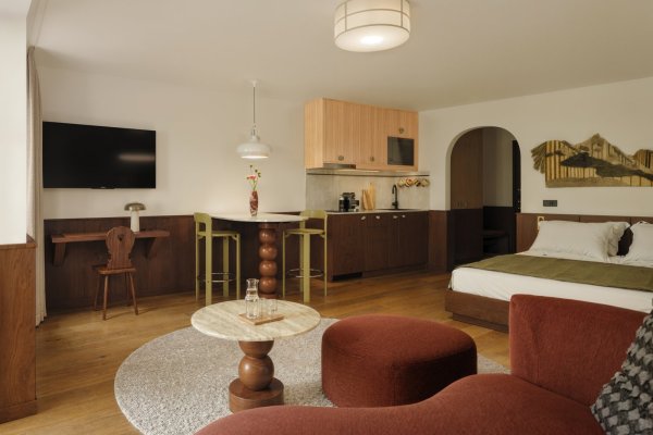 Der Raum zeigt ein Bett, einen Tisch, einen Fernseher und eine kleine Küche. Es gibt einen runden Teppich auf dem Boden.