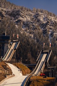 Zwei Skisprungschanzen stehen in den Alpen. Schneebedeckte Bäume umgeben die Schanzen. Es ist ein klarer Wintertag.
