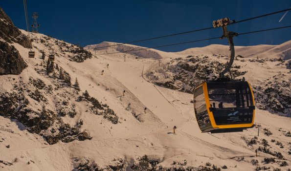 Seilbahn fährt über eine Skipiste im Gebirge Eine Seilbahn transportiert Skifahrer über eine schneebedeckte Piste in den Bergen. Es ist sonnig und die Leute genießen die Abfahrt.