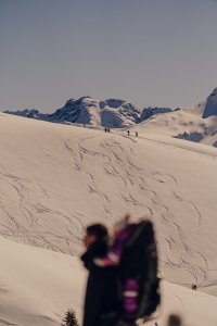 Einige Menschen gehen auf einem verschneiten Hügel in den Bergen. Im Hintergrund sind hohe Gipfel sichtbar.