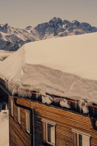 Eine Berghütte steht unter einem schneebedeckten Dach. Die Umgebung zeigt Berge im Hintergrund unter blauem Himmel.
