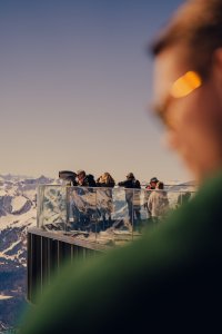 Menschen stehen auf einer Aussichtsplattform und betrachten die Berge. Es ist ein klarer Tag mit schneebedeckten Gipfeln.