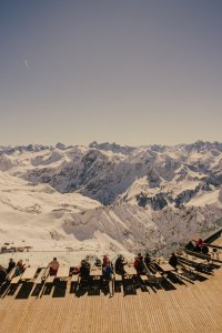 Menschen sitzen auf einer Terrasse und schauen auf die Berge. Es ist sonnig und viele Schneefelder sind sichtbar.
