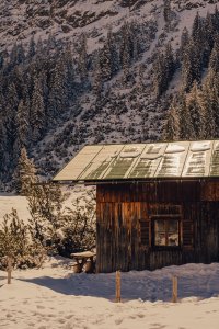 Die Hütte steht im Schnee zwischen Bäumen. Die Sonne scheint auf das Dach und es ist Winter.