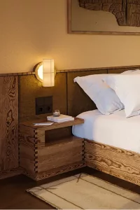 Das Zimmer zeigt ein Holzbett mit weißen Kissen, einem Nachttisch und einer Lampe an der Wand. Es ist Hell.