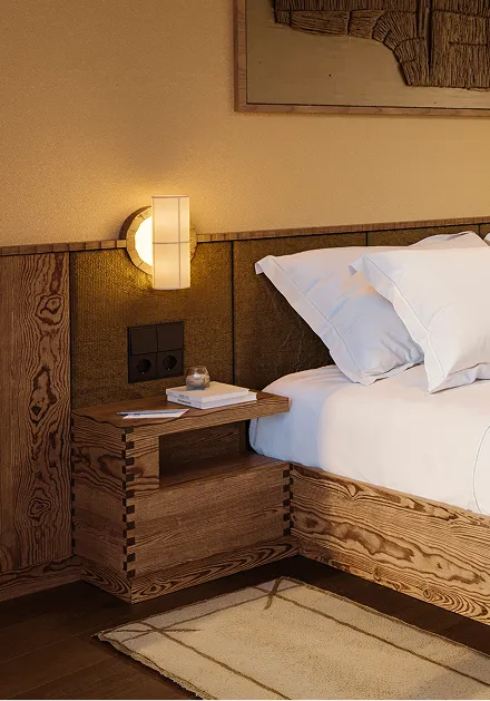 Das Zimmer zeigt ein Holzbett mit weißen Kissen, einem Nachttisch und einer Lampe an der Wand. Es ist Hell.