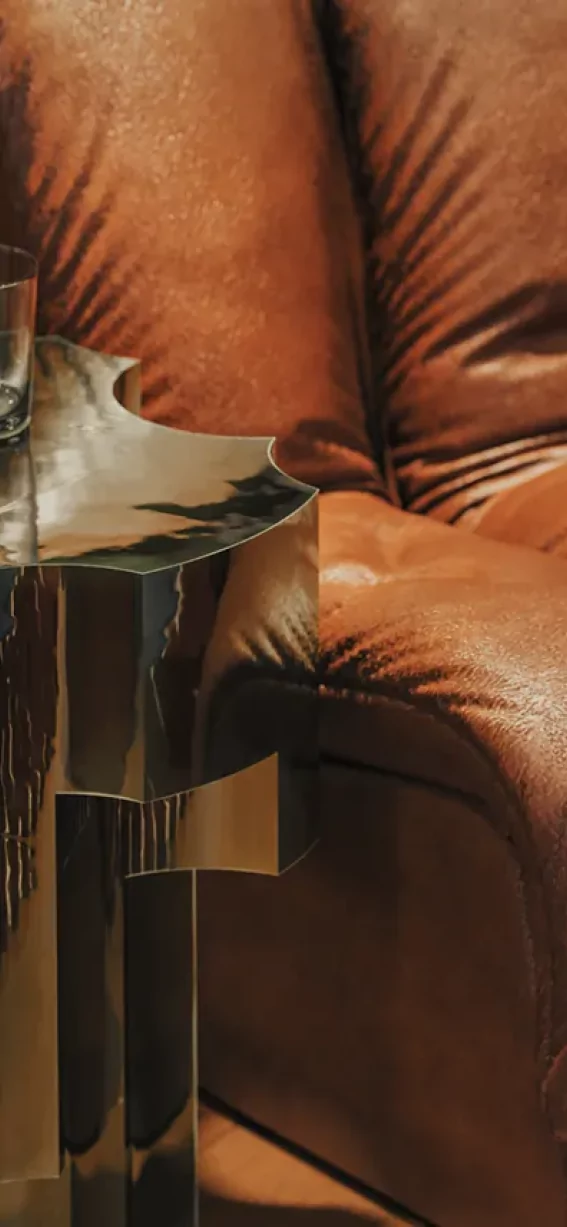In einem Raum steht ein braunes Sofa neben einem Tisch. Auf dem Tisch steht ein Glas. Es ist Abend und es gibt warmes Licht.