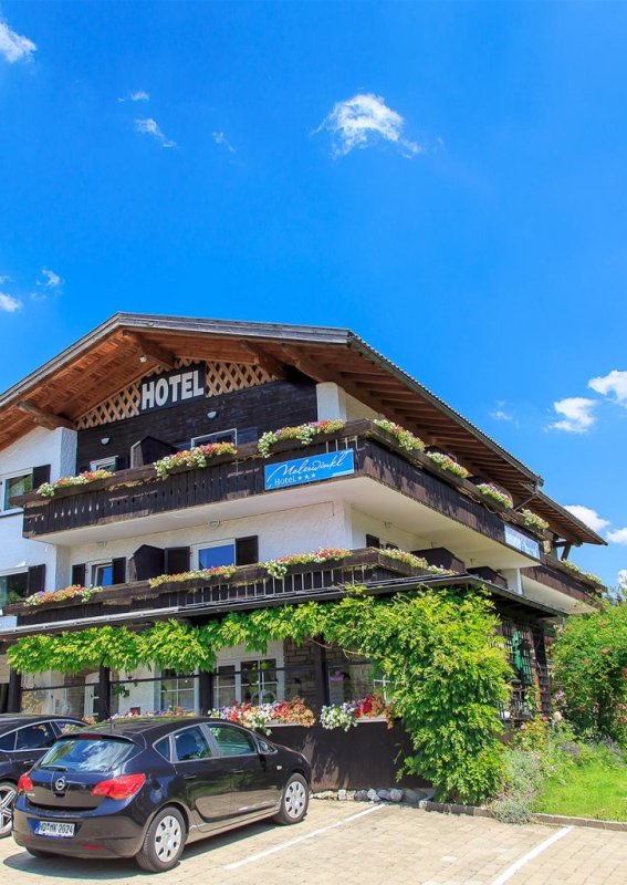 Das Hotel steht in einer ländlichen Umgebung. Es gibt viele Autos auf dem Parkplatz. Die Sonne scheint, der Himmel ist blau.