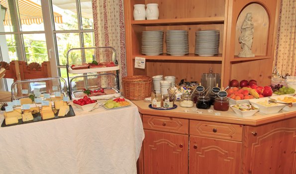 Es gibt viele Speisen auf dem Buffet. Käse, Obst und Brot stehen bereit. Die Sonne scheint ins Zimmer.