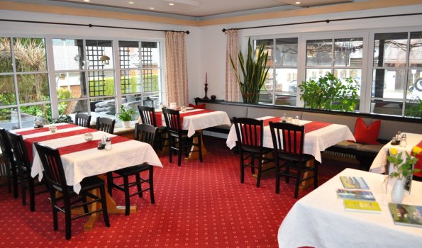 Im Restaurant stehen Tische mit weißen Tischtüchern. Die Fenster zeigen Pflanzen und eine grüne Aussicht.