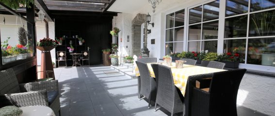 Auf der Terrasse steht ein Tisch mit Stühlen. Pflanzen sind um den Bereich angeordnet. Es ist ein sonniger Tag.