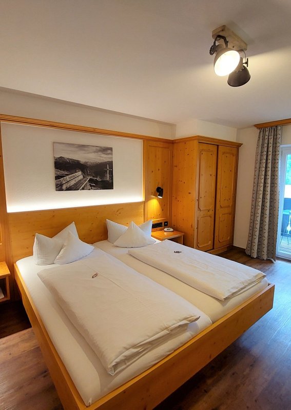 Rustikales Zimmer in einer Berghütte Das Zimmer hat zwei Betten und einen Balkon mit Blick auf die Berge. Es gibt Holzverkleidungen und einfache Möbel.