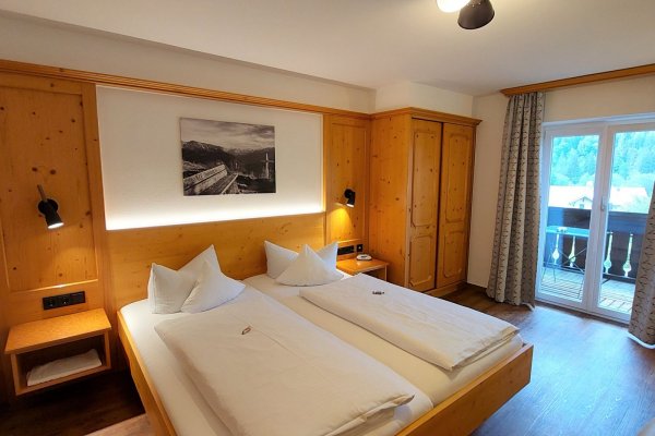 Rustikales Zimmer in einer Berghütte Das Zimmer hat zwei Betten und einen Balkon mit Blick auf die Berge. Es gibt Holzverkleidungen und einfache Möbel.