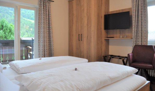 In einem Zimmer mit zwei Betten sind die Fenster geöffnet. Das Zimmer zeigt den Blick auf die Berge und den Balkon.