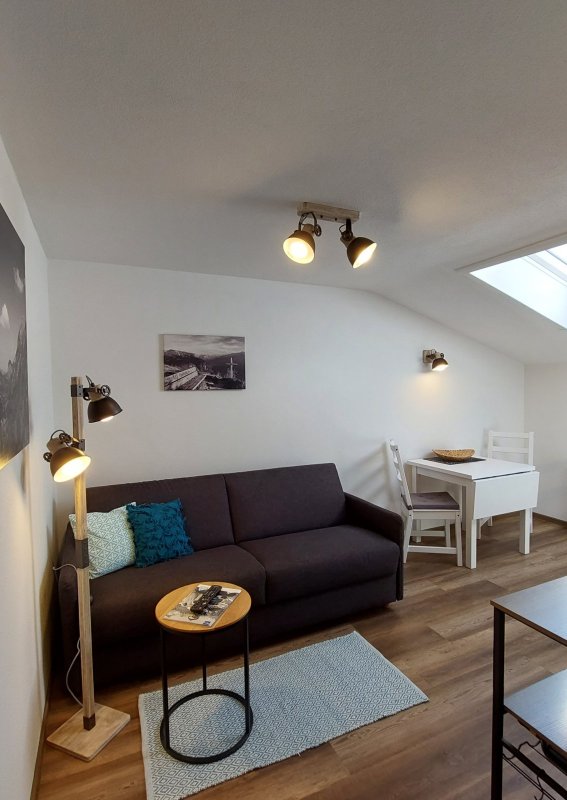 In einem kleinen Raum steht ein Sofa und ein Tisch. Ein Fenster lässt Licht hereinkommen und die Wände sind hell.