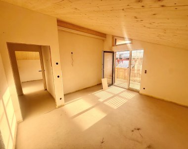 Das Zimmer zeigt eine leere Fläche mit Fenstern und einem Balkon. Der Raum wirkt hell und hat einen Holzfußboden.