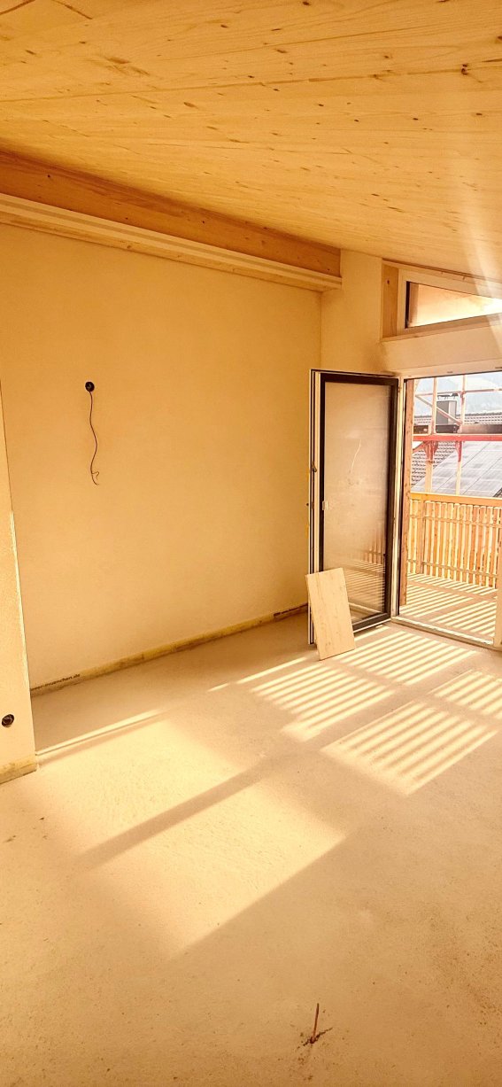 Das Zimmer zeigt eine leere Fläche mit Fenstern und einem Balkon. Der Raum wirkt hell und hat einen Holzfußboden.
