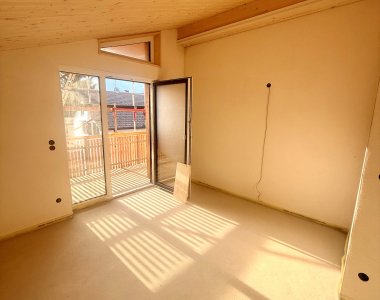 Der Raum hat einen großen Fenster mit Licht. Der Boden ist beige und unmöbliert. Sonnenstrahlen fallen ins Zimmer.