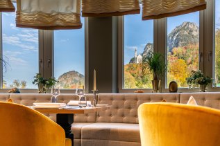 Einblick in das Restaurant des AMERON Neuschwanstein Hotels mit gedeckten Tischen und Ausblick auf Schloss Neuschwanstein im Herbst.