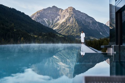 Ein Mann steht am Rande des Außenpools im Travel Charme Ifen Hotel im Kleinwalsertal und hinter im erstrecken sich die Berge