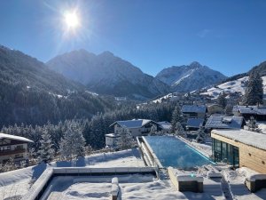 Die Außenansicht des verschneiten Travel Charme Ifen Hotels im Kleinwalsertal mit Infinitypool und Bergpanorama.