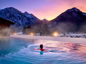 Eine Frau schwimmt im Außenpool des Hotel Prinz-Luitpold-Bad in Bad Hindelang im Winter und genießt einen Sonnenuntergang und das Bergpanorama