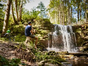 Ein Mann unternimmt eine Radtour durch die Umgebung des Hanusel Hof in Weitnau und hält für eine Pause an einem Wasserfall
