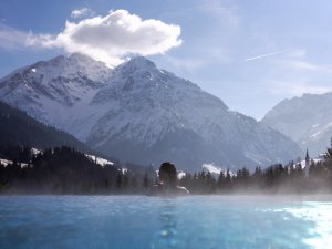 Ein Gast im Travel Charme Ifen Hotel in Kleinwalsertal schwimmt im Außenpool und genießt die Aussicht