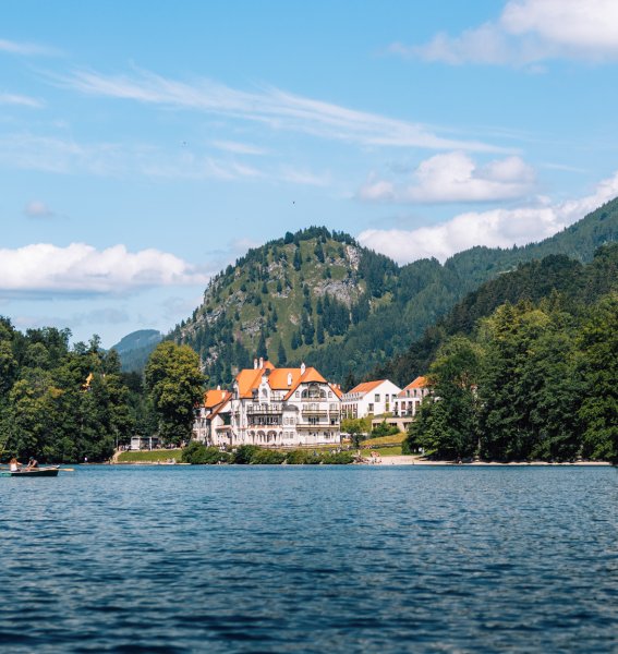 Ansicht der Hotel AMERON Alpsee Resort und Spa in Hohenschwangau vor einer Bergkulisse und den umliegenden Schlössern. Im Vordergrund ist ein See.