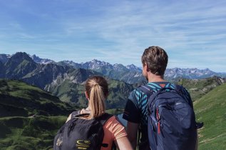 Ansicht einer Frau und eines Mannes, die eine Wanderung in den Bergen machen. Im Hintergrund ist eine Bergkulisse zu sehen.