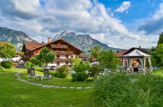 Außenansicht des Hotel Helmer in Schwangau und dessen Garten im Sommer, im Hintergrund ist eine Bergkulisse zu sehen.
