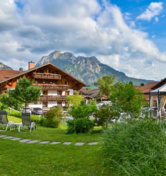 Außenansicht des Hotel Helmer in Schwangau und dessen Garten im Sommer, im Hintergrund ist eine Bergkulisse zu sehen.
