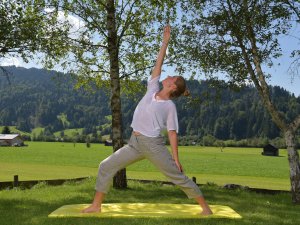 Eine Frau macht eine Yogaübung auf der Wiese im Außenbereich des Hotels Alpenhof vor Bergpanorama im Sommer.
