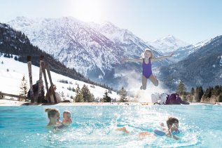 Eine Familie hat im Außenpool des Oberjoch - Familux Resort Spaß während hinter ihr die verschneiten Berge zu sehen sind