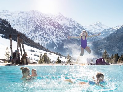 Eine Familie hat im Außenpool des Oberjoch - Familux Resort Spaß während hinter ihr die verschneiten Berge zu sehen sind