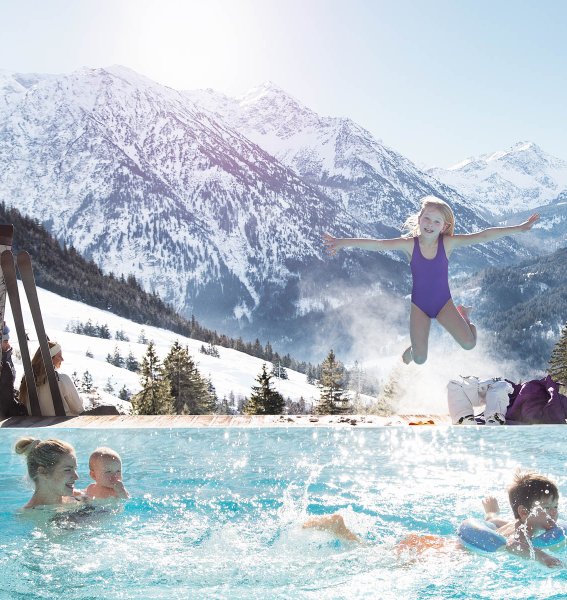 Eine Familie hat im Außenpool des Oberjoch - Familux Resort Spaß während hinter ihr die verschneiten Berge zu sehen sind
