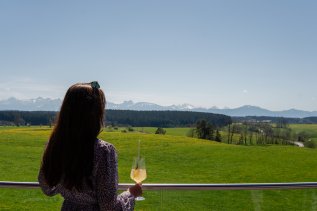 Eine Frau steht mit einem Cocktail am Gelände auf der Terrasse im Das Weitblick Allgäu in Marktoberdorf und genießt die Aussicht auf grüne Wiesen