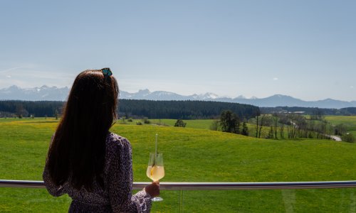 Das Weitblick Allgäu Aussicht Sommer Eine Frau steht mit einem Cocktail am Gelände auf der Terrasse im Das Weitblick Allgäu in Marktoberdorf und genießt die Aussicht auf grüne Wiesen