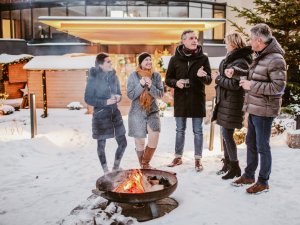 Personen stehen gemeinsam vor dem Bergkristall - Mein Resort im Allgäu in Oberstaufen im Schnee vor einer Feuerschale und unterhalten sich