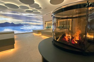 Neuer Ruheraum im Wellnessbereich des Bayerischen Hofs in Oberstaufen mit Blick auf ein Bergpanoramabild und einen Kamin mit Feuer.