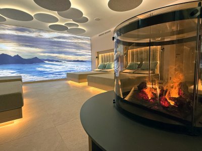 Neuer Ruheraum im Wellnessbereich des Bayerischen Hofs in Oberstaufen mit Blick auf ein Bergpanoramabild und einen Kamin mit Feuer.