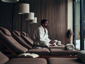 Ein Mann sitzt im Bademantel auf einer der Liegen im Ruheraum im Ifen Hotel Kleinwalsertal und hört Musik während er aus den bodentiefen Fenstern schaut