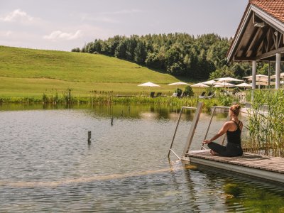 Eine Frau macht am See vom Haubers Naturresort in Oberstaufen umgeben von grünen Wiesen Yoga