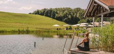 Eine Frau macht am See vom Haubers Naturresort in Oberstaufen umgeben von grünen Wiesen Yoga