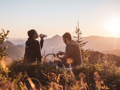 Zwei Personen sitzen nach einer Wanderungen auf dem Gipfel und genießen die Aussicht während sie aus Allgäuer Alpenwasser Flaschen trinken