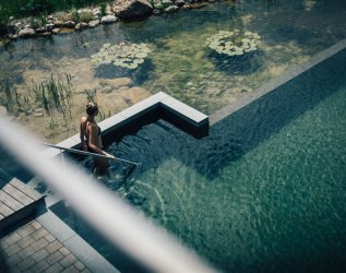 Blick von oben auf den natürlichen Außenpool des Hotels Torghele's Wald & Fluh in Balderschwang. Frau betritt den Pool.
