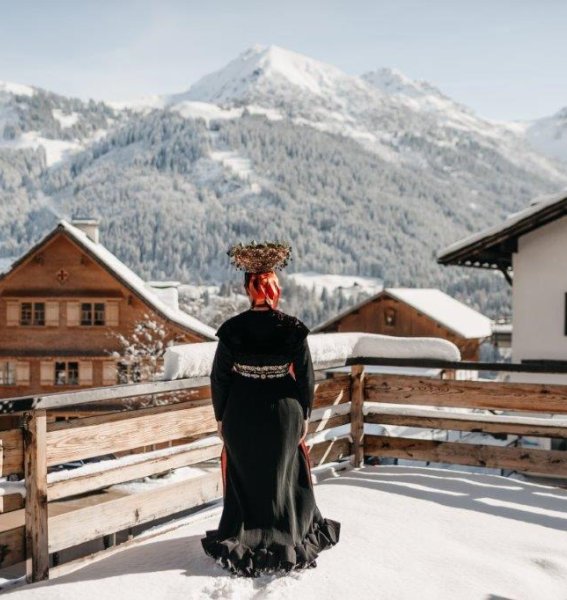 Eine Dame in einer traditionellen Tracht steht auf einem schneebedeckten Balkon im Hotel Tradizio im Kleinwalsertal.