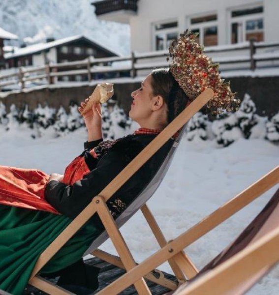 Eine Dame in traditioneller Tracht genießt ein Eis auf einer Liege im schneebedeckten Garten im Hotel Tradizio im Kleinwalsertal.