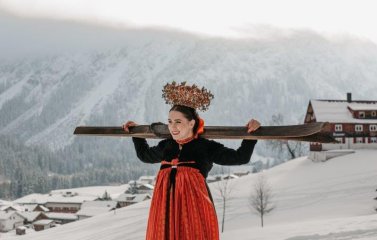 Eine Dame in traditioneller Tracht hält ein paar Ski über ihren Schultern im Schnee in der Nähe des Hotels Tradizio im Kleinwalsertal.