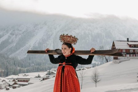 Eine Dame in traditioneller Tracht hält ein paar Ski über ihren Schultern im Schnee in der Nähe des Hotels Tradizio im Kleinwalsertal.